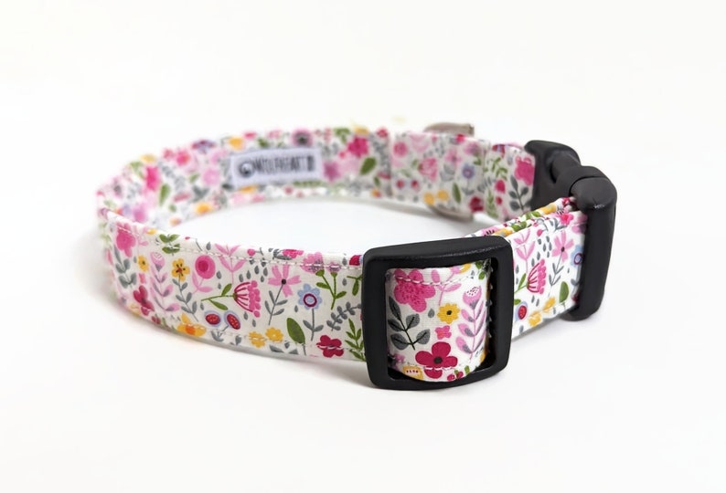 May include: A white dog collar with a floral print featuring pink, yellow, and blue flowers. The collar has a black plastic buckle.