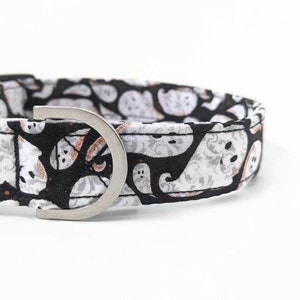 May include: A Halloween-themed dog collar featuring a black background with white and gray ghost patterns. The collar has a silver-colored metal buckle and a sturdy design, perfect for pet owners looking for festive accessories.