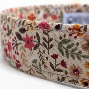 May include: A beige fabric dog collar with a floral pattern featuring pink, red, brown, and yellow flowers. The collar has a stitched edge.