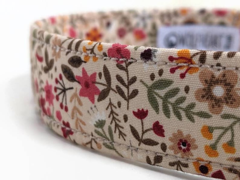 May include: A beige fabric dog collar with a floral pattern featuring pink, red, brown, and yellow flowers. The collar has a stitched edge.