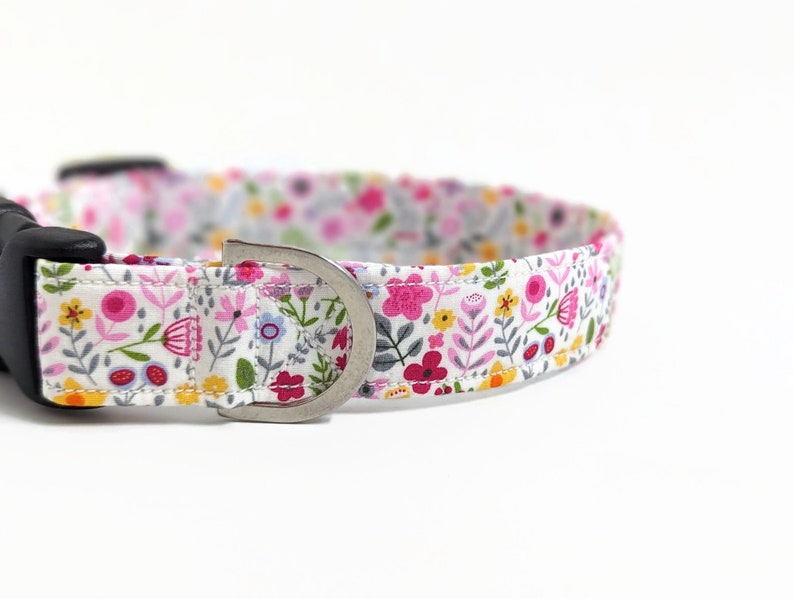 May include: A white dog collar with a floral print featuring pink, yellow, and green flowers. The collar has a black buckle and a silver D-ring.