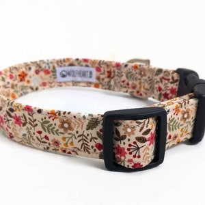 May include: A beige dog collar with a floral pattern featuring red, orange, yellow, and green flowers. The collar has a black buckle and the text "Wildheart" is printed on the collar.