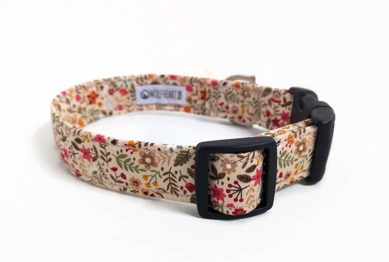 May include: A beige dog collar with a floral pattern featuring red, orange, yellow, and green flowers. The collar has a black buckle and the text "Wildheart" is printed on the collar.