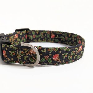 May include: A black dog collar with a festive pattern featuring red birds, green holly leaves, and gold stars. The collar has a black plastic buckle and a silver metal D-ring for leash attachment.