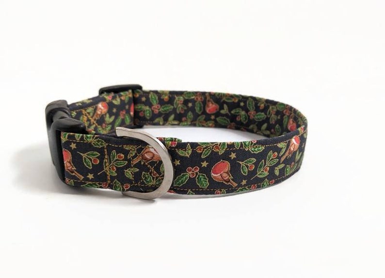 May include: A black dog collar with a festive pattern featuring red birds, green holly leaves, and gold stars. The collar has a black plastic buckle and a silver metal D-ring for leash attachment.