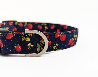 May include: A dark blue fabric pet collar featuring a repeating pattern of red strawberries, green leaves, and small white dots. The collar includes a silver-toned metal D-ring and a black plastic side-release buckle for secure fastening.