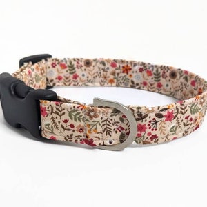 May include: A beige dog collar with a floral pattern featuring red, orange, and yellow flowers and green leaves. The collar has a black plastic buckle and a silver metal D-ring.
