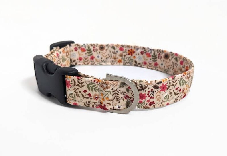 May include: A beige dog collar with a floral pattern featuring red, orange, and yellow flowers and green leaves. The collar has a black plastic buckle and a silver metal D-ring.