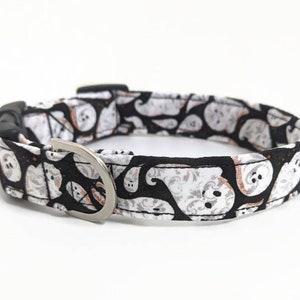 May include: A Halloween-themed dog collar featuring a black background with white and orange ghost patterns. The collar has a silver-colored metal D-ring and a black plastic buckle. The fabric is patterned with various ghost designs.