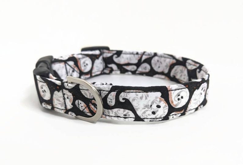 May include: A Halloween-themed dog collar featuring a black background with white and orange ghost patterns. The collar has a silver-colored metal D-ring and a black plastic buckle. The fabric is patterned with various ghost designs.