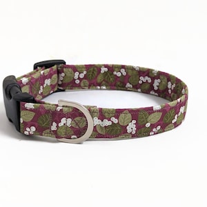 May include: A burgundy dog collar with a white and green floral pattern. The collar has a black plastic buckle and a silver metal D-ring.