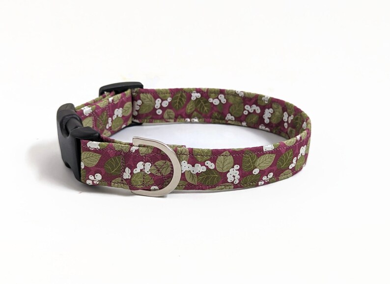 May include: A burgundy dog collar with a white and green floral pattern. The collar has a black plastic buckle and a silver metal D-ring.