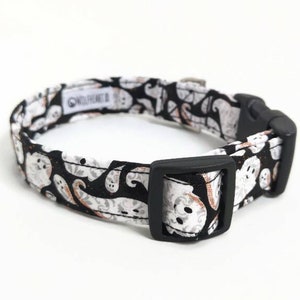 May include: A black and white dog collar with a repeating ghost pattern. The collar has a black plastic buckle and a metal loop for leash attachment. The fabric features white ghosts on a black background.