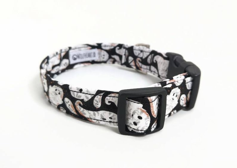 May include: A black and white dog collar with a repeating ghost pattern. The collar has a black plastic buckle and a metal loop for leash attachment. The fabric features white ghosts on a black background.