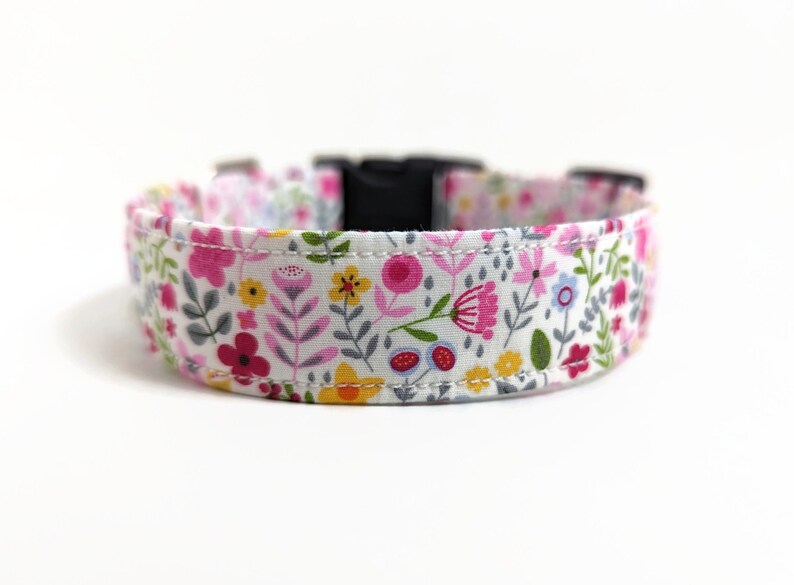 May include: White dog collar with a floral print featuring pink, yellow, and green flowers. The collar has a black plastic buckle.