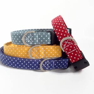 May include: A stack of four fabric pet collars featuring a white polka dot pattern on red, blue, yellow, and grey backgrounds. Each collar includes a silver metal D-ring for lead attachment and a black plastic side-release buckle for secure fastening.