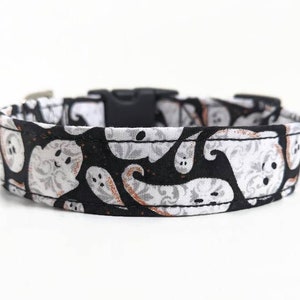 May include: Black and white Halloween-themed dog collar featuring a repeating pattern of friendly ghost figures. The collar has a black plastic buckle and a metal loop for leash attachment. The fabric design includes gray floral accents.
