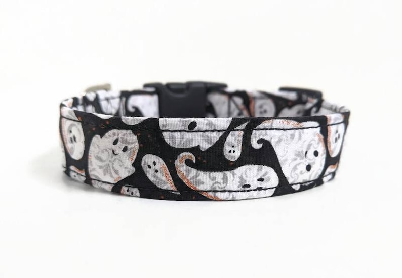 May include: Black and white Halloween-themed dog collar featuring a repeating pattern of friendly ghost figures. The collar has a black plastic buckle and a metal loop for leash attachment. The fabric design includes gray floral accents.