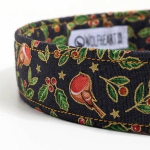 May include: Close-up of a dog collar with a festive design. The collar features a dark background with gold accents, and illustrations of robins, holly, and stars. The brand name "WOLFHEART" is visible on a white tag.