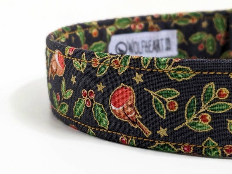 May include: Close-up of a dog collar with a festive design. The collar features a dark background with gold accents, and illustrations of robins, holly, and stars. The brand name "WOLFHEART" is visible on a white tag.
