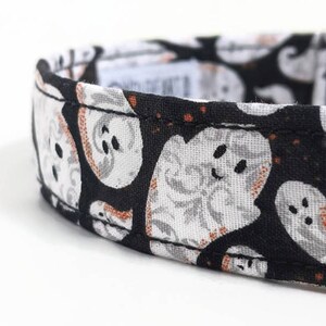 May include: Close-up of a black and white Halloween-themed collar. The collar features a repeating pattern of smiling ghost faces with orange accents. The collar is made of fabric and has a stitched edge.