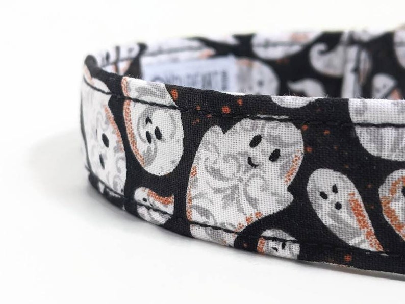 May include: Close-up of a black and white Halloween-themed collar. The collar features a repeating pattern of smiling ghost faces with orange accents. The collar is made of fabric and has a stitched edge.