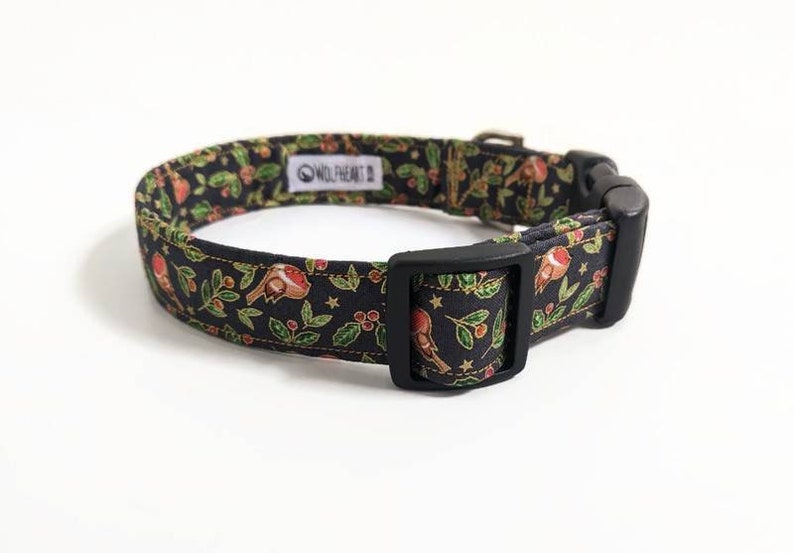 May include: Black dog collar with a festive pattern of birds, berries, and leaves. The collar has a black plastic buckle and a metal adjustment piece. The brand name "WOLFHEART" is visible on a white tag.