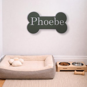 May include: A dog bed with a plush, beige interior and a grey exterior, alongside a wooden dog bowl stand with two stainless steel bowls. A bone-shaped sign with the name "Phoebe" hangs above.