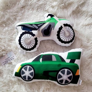 May include: Two plush toys shaped like a green dirt bike and a green sports car. The toys are on a white fluffy surface.