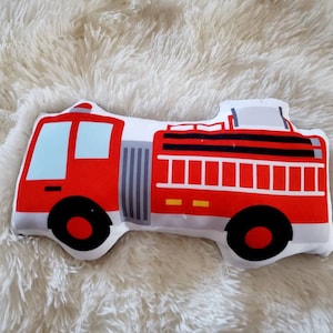 May include: A red and white fire truck shaped pillow with black wheels and blue windows. The pillow is designed with a detailed illustration of a fire truck, perfect for a child's room or nursery.