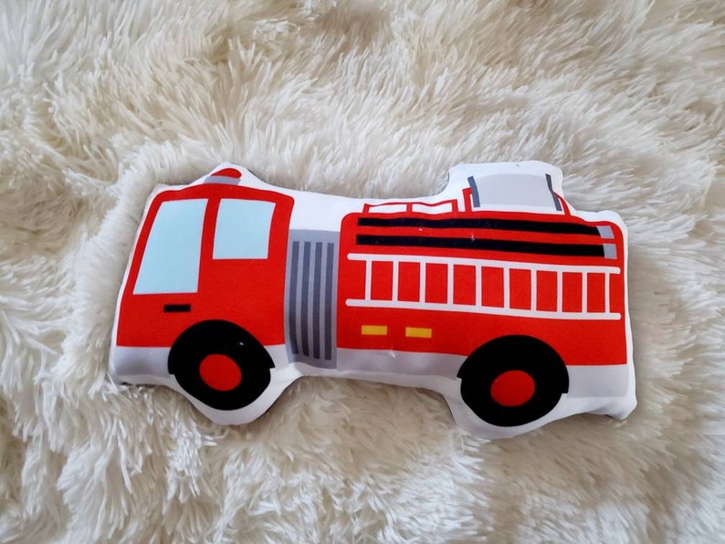 May include: A red and white fire truck shaped pillow with black wheels and blue windows. The pillow is designed with a detailed illustration of a fire truck, perfect for a child's room or nursery.