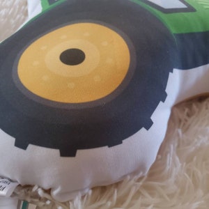 May include: Close-up of a green and black tractor cushion with a yellow tyre. The cushion is made of soft fabric and has a white background.