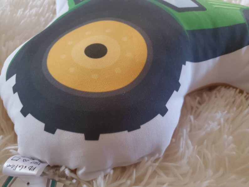 May include: Close-up of a green and black tractor cushion with a yellow tyre. The cushion is made of soft fabric and has a white background.