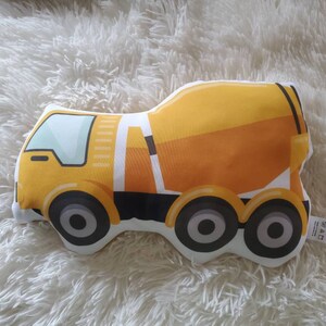 May include: A yellow and orange cement truck plush toy with black wheels and a white outline. The toy is designed with a cartoonish style and is set against a fluffy white background.