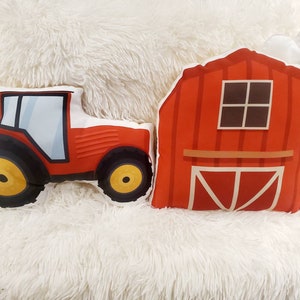 May include: Two red plush pillows shaped like a tractor and a barn. The tractor has black wheels with a yellow centre. The barn has a white cloud on top and a brown window.
