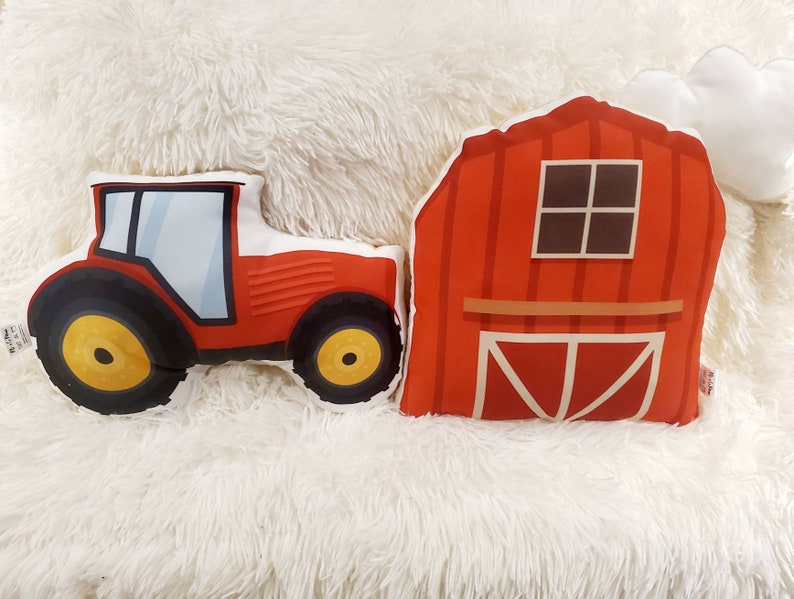 May include: Two red plush pillows shaped like a tractor and a barn. The tractor has black wheels with a yellow centre. The barn has a white cloud on top and a brown window.