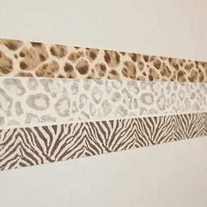 May include: Three horizontal strips of adhesive tape arranged in a stack. The top strip features a brown and tan leopard print. The middle strip displays a light gray and white leopard pattern. The bottom strip shows a brown and white zebra stripe design.