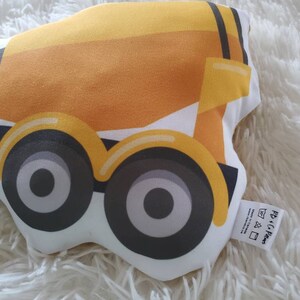 May include: A yellow and white construction truck-shaped pillow with black and gray wheels. The pillow has a tag that reads "PB + C Paws". The pillow is on a white, fluffy surface.