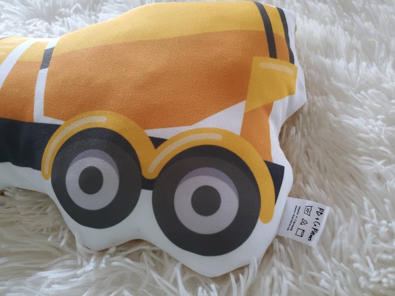 May include: A yellow and white construction truck-shaped pillow with black and gray wheels. The pillow has a tag that reads "PB + C Paws". The pillow is on a white, fluffy surface.