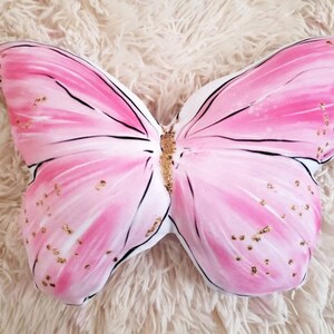 Puede incluir: Una almohada en forma de mariposa rosa con detalles de purpurina dorada. La almohada est&aacute; hecha de tela suave y tiene un fondo blanco.