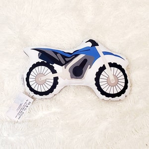 May include: A blue and gray plush pillow shaped like a dirt bike. The pillow has a white background and black wheels.