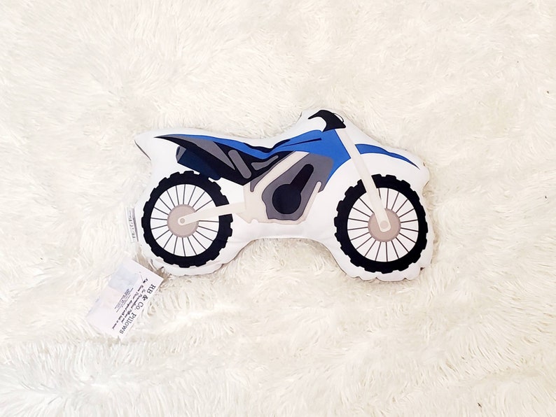 May include: A blue and gray plush pillow shaped like a dirt bike. The pillow has a white background and black wheels.