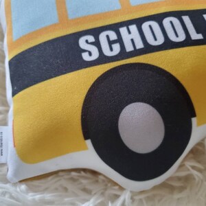 May include: A yellow school bus-shaped pillow with black tires and the words "SCHOOL BUS" in white. The pillow has a blue sky design at the top and a white border. A small tag with text is attached to the side.