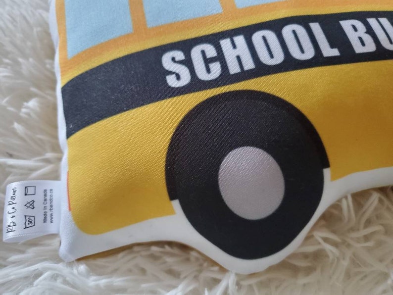 May include: A yellow school bus-shaped pillow with black tires and the words "SCHOOL BUS" in white. The pillow has a blue sky design at the top and a white border. A small tag with text is attached to the side.
