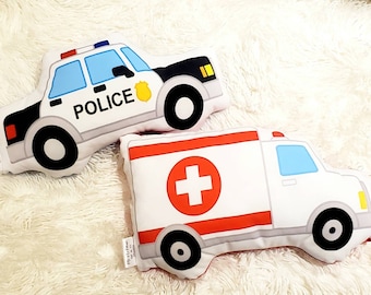 May include: Two plush toy pillows shaped like vehicles. The first pillow is a blue and black police car with the word "POLICE" printed on the side. The second pillow is a white ambulance with a red cross on the side.
