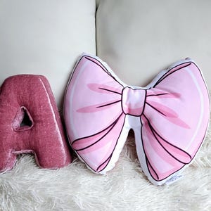 Peut inclure: Deux coussins décoratifs, l'un en forme de lettre A de couleur rose foncé et l'autre en forme de nœud rose.