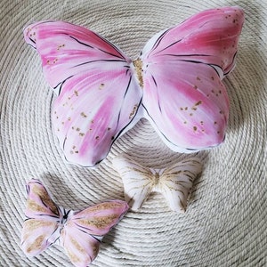 Puede incluir: Tres almohadas en forma de mariposa de color rosa y blanco con detalles de purpurina dorada. Las almohadas est&aacute;n sobre una superficie tejida de color crema.