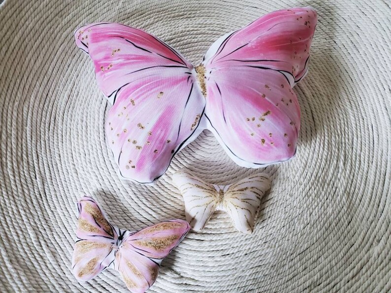 Puede incluir: Tres almohadas en forma de mariposa de color rosa y blanco con detalles de purpurina dorada. Las almohadas est&aacute;n sobre una superficie tejida de color crema.