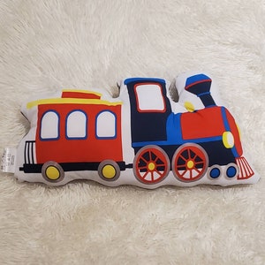 May include: A red, blue, and yellow train pillow with a white background. The train has three cars, including a caboose, a passenger car, and an engine.