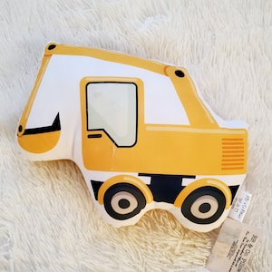 May include: A yellow and white excavator-shaped cushion with black wheels and accents. The cushion has a window and a black scoop. The cushion is on a white, fluffy surface. A tag is attached to the cushion.